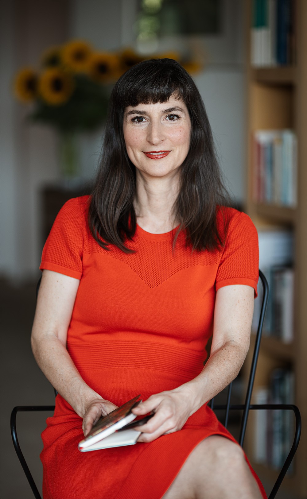 Porträtfoto-Dr.-Freya-Amann-auf-einem-Stuhl-sitzend-mit-einem-Buch-in-der-Hand-im-Hintergrund-befindet-sich-ein-ruhig-gestalteter-Innenraum-mit-Sonnenblumenstrauß-Bild-und-Bücherschrank